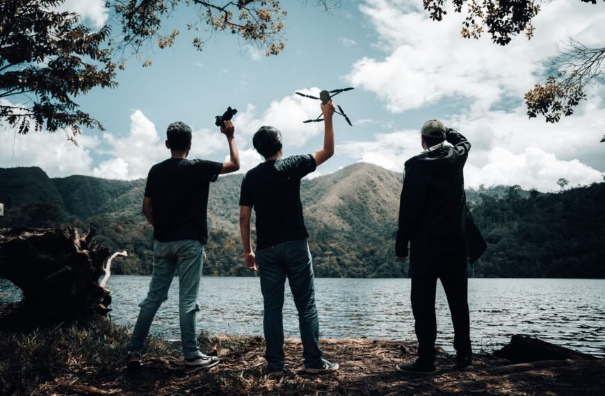 Three men holding drones by a mountain lake, silhouette against a scenic backdrop.